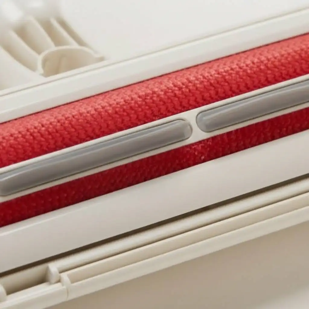 Macro diagonal view of a white pet hair remover roller featuring textured red velvet strips and a grey silicone squeegee blade designed to trap fur. Macro diagonal view of a white pet hair remover roller featuring textured red velvet strips and a grey silicone squeegee blade designed to trap fur.